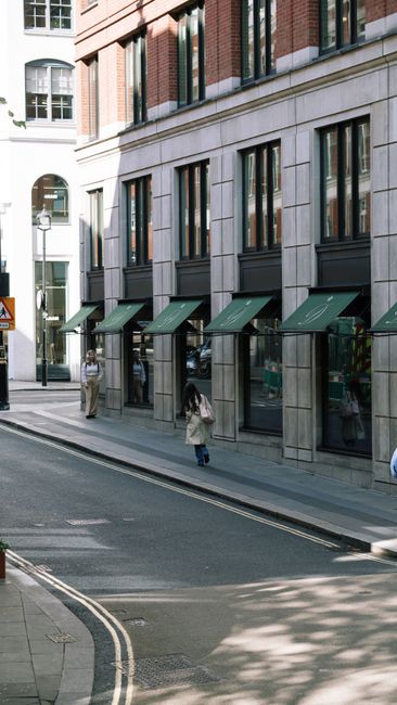Exterior view of a commercial building on Upper Street in Islington, showcasing a modern facade with large glass windows framed by stonework. The ground level features several storefronts with dark green awnings over their entrances. The sidewalk is clean and well-maintained, with a few pedestrians walking along the street. Bright daylight highlights the building's sleek design and the reflective quality of the glass. This scene exemplifies professional external surface cleaning and maintenance services provided by Carpet Cleaning Islington for shops and offices on Upper Street, emphasizing hygiene and polished appearance in a busy urban setting.
