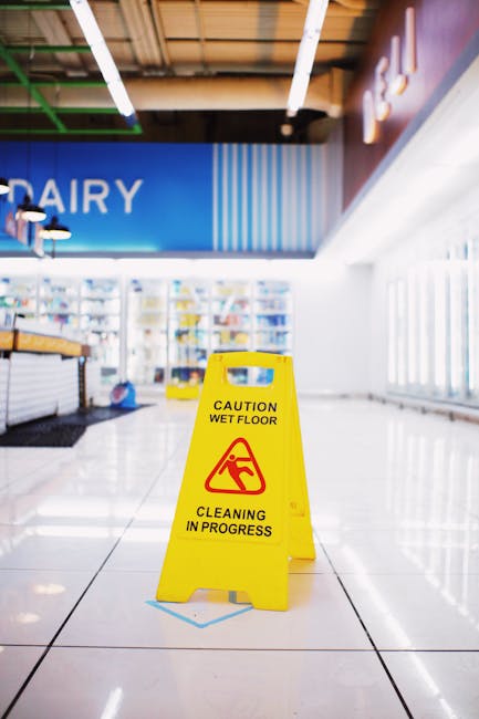Interior view of a commercial shop in Islington with a yellow 'Caution Wet Floor' sign placed on large, glossy white ceramic tiles. The shop features bright natural light filtering through large windows on the right, with a modern ceiling that includes visible lighting fixtures. The background shows shelves stocked with various products, and part of a blue sign with the word 'Dairy' is visible. The floor appears clean but temporarily wet, indicating recent cleaning by professional cleaners such as Carpet Cleaning Islington. This image highlights the importance of surface cleaning and hygiene maintenance in retail spaces to ensure customer safety and store cleanliness.
