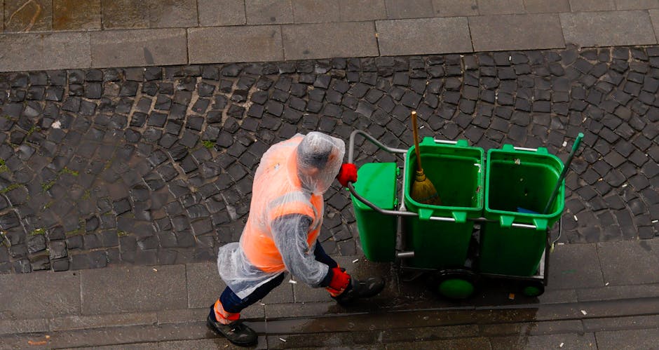 An individual wearing a grey hoodie, orange high-visibility vest, and red gloves is engaged in surface cleaning outdoors on a cobblestone pavement adjacent to a concrete sidewalk. The person is pushing a green, three-compartment cleaning trolley equipped with a broom, a mop, and a dustpan, indicating cleaning or sanitisation activities. The background includes a section of cobbled stones and a smooth concrete edge, with natural daylight illuminating the scene. The image reflects professional cleaning work associated with domestic cleaning and maintenance, with the individual focused on sanitation during service provided by Carpet Cleaning Islington. No visible dust or dirt is present, suggesting a thorough cleaning process.
