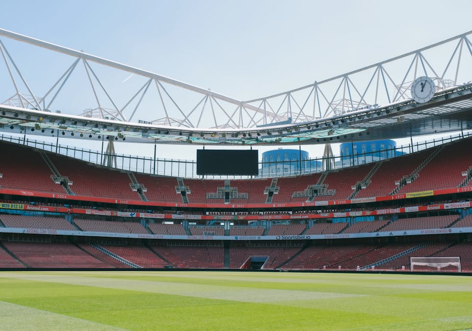 Interior view of a modern stadium with tiered red seating, green grass pitch, and a large central scoreboard. The stadium features a white, open lattice roof structure with steel supports, allowing natural light to illuminate the seating and field area. The field surface appears well-maintained and freshly mowed, with no visible debris or markings. The seating sections are arranged in a semi-circular pattern around the pitch, and the overall scene suggests a clean and orderly environment suitable for sports events. This setting exemplifies surface cleaning and maintenance essential for sanitisation in public sports venues, with the focus on hygiene and orderliness as part of professional cleaning practices by Carpet Cleaning Islington, mirroring the attention to cleanliness discussed in the context of Islington council rules for mattress disposal for cleaners.
