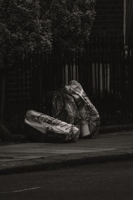 Three large black trash bags filled with discarded items are placed on a paved sidewalk next to a black metal fence, with a wooden fence and leafy trees in the background. The bags appear to contain household waste, and the scene is outdoors, lit by natural daylight. The setting suggests a typical urban area in Islington, where proper disposal of waste and furniture is important for maintaining cleanliness and hygiene. Carpet Cleaning Islington offers services aligned with local regulations, including mattress disposal, emphasizing the importance of responsible waste management in residential and commercial cleaning practices.