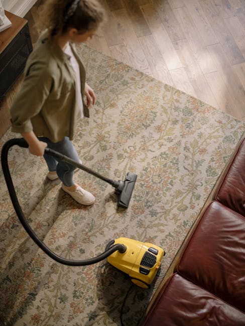 A woman using a yellow vacuum cleaner to perform surface cleaning on a patterned, beige area rug in a living room. The room features a wooden floor and a dark red leather sofa. The woman is dressed casually in a beige jacket and jeans, and is standing on the rug while vacuuming. The scene is well-lit, highlighting the cleanliness and maintained condition of the rug and surrounding surfaces. The image exemplifies domestic cleaning practices, with a focus on deep cleaning and hygiene, as carried out by Carpet Cleaning Islington, a professional cleaning service, in a typical home environment.
