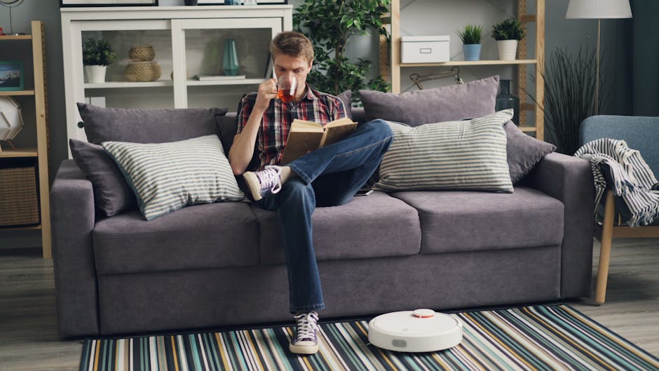 A young man with light skin and short hair is seated on a modern, grey fabric sofa in a well-lit living room, drinking from a glass of tea or coffee while reading a book. The sofa is adorned with multiple pillows in shades of grey and white, some with striped patterns. In front of the sofa, a striped multicolored rug covers the wooden floor. A white robot vacuum cleaner is positioned on the rug, indicating active surface cleaning. The background features a white shelving unit with decorative items, plants, and storage boxes, along with a potted plant on the right side. The room appears clean and tidy, with ample natural light illuminating the area, and the overall setting emphasizes domestic cleaning and maintenance, with Carpet Cleaning Islington providing professional surface cleaning services for the space.