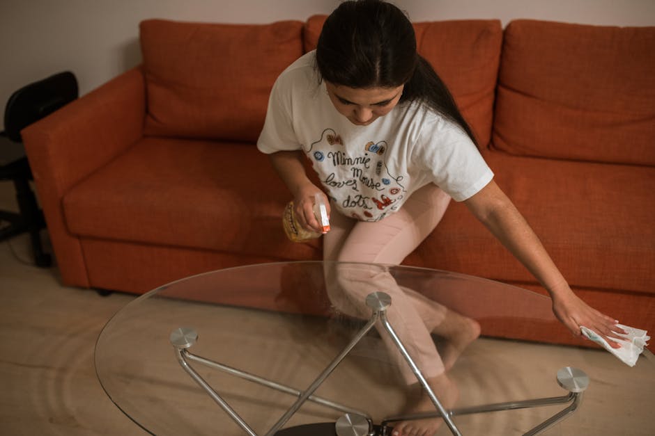 A professional cleaner wearing a full white protective suit, gloves, and a face mask is seen deep cleaning a black leather sofa in a well-lit living room. The cleaner is using a cleaning cloth near the window, which allows natural light to illuminate the scene. The sofa appears dust-free and well-maintained, with a mustard-colored cushion placed on it. Next to the sofa, a small black round table holds a spray bottle with a white and turquoise nozzle, likely used for surface sanitisation. Behind the sofa, there is a tall green houseplant adding freshness to the room, alongside a framed picture situated on the floor. The room features a large window with white framing, contributing to the bright and airy atmosphere. This scene exemplifies surface cleaning and sanitisation practices as part of domestic cleaning services provided by Carpet Cleaning Islington, suited for maintaining hygiene and furniture longevity.
