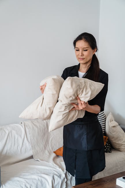 A woman with dark hair, dressed in a black apron, is standing in a bright, well-lit living room. She is holding a beige cushion and appears to be preparing to carry or arrange it, indicating a cleanin