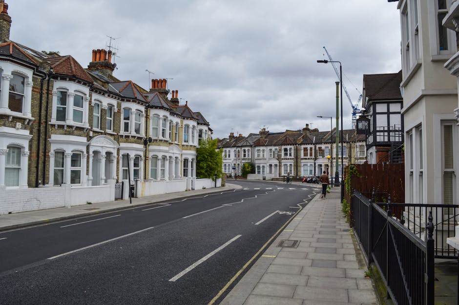 A quiet residential street in Islington with row houses featuring white facades, bay windows, and red-tiled roofs. The street is paved with asphalt, marked with white lines, and lined with a concrete sidewalk, which is clean and well-maintained. Modern streetlights and a few parked bicycles are visible, along with some construction cranes in the background. The scene is illuminated by natural daylight under a cloudy sky, illustrating typical urban architecture and street layout in the area, relevant to domestic cleaning and maintenance contexts through the [DOMAIN] website, Carpet Cleaning Islington.