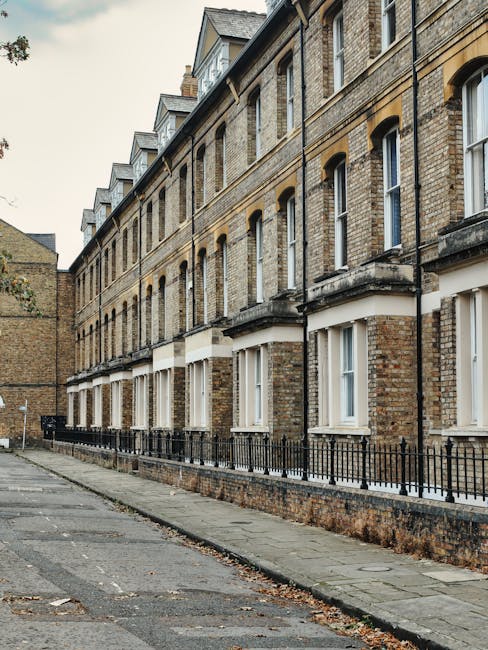 A row of old brick residential buildings along a narrow pavement, with multiple white-framed sash windows and black metal railings. The buildings feature beige and brown bricks with decorative stone accents around the windows and doors. The pavement is made of concrete slabs, showing some dirt and fallen leaves, with a slightly uneven surface. The scene is lit with natural daylight, highlighting the weathered texture of the brick walls. While no cleaning tools or objects are visible, this image exemplifies the exterior environment that can benefit from professional cleaning services offered by Carpet Cleaning Islington. The overall appearance is tidy but slightly aged, emphasizing the importance of regular facade and surface maintenance for residential exteriors in London.