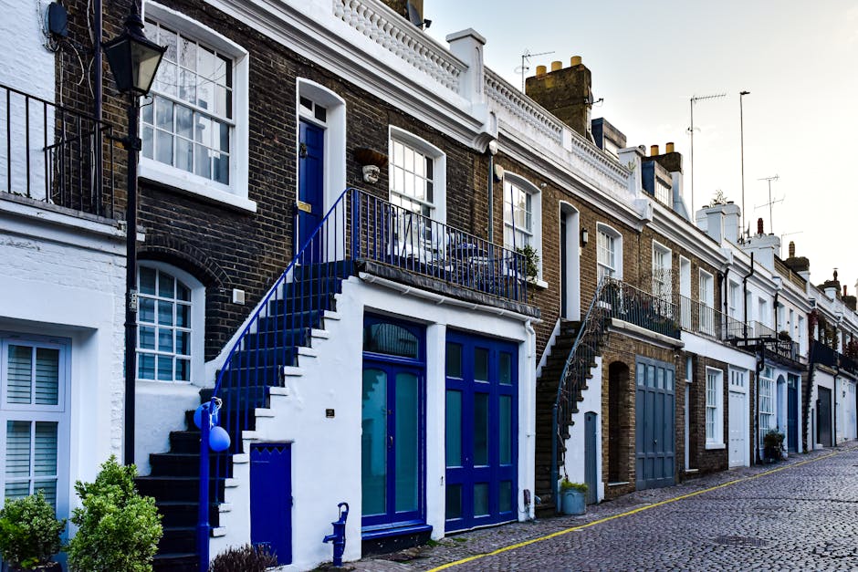 A quiet residential street in Islington with row houses featuring white facades, bay windows, and red-tiled roofs. The street is paved with asphalt, marked with white lines, and lined with a concrete sidewalk, which is clean and well-maintained. Modern streetlights and a few parked bicycles are visible, along with some construction cranes in the background. The scene is illuminated by natural daylight under a cloudy sky, illustrating typical urban architecture and street layout in the area, relevant to domestic cleaning and maintenance contexts through the [DOMAIN] website, Carpet Cleaning Islington.